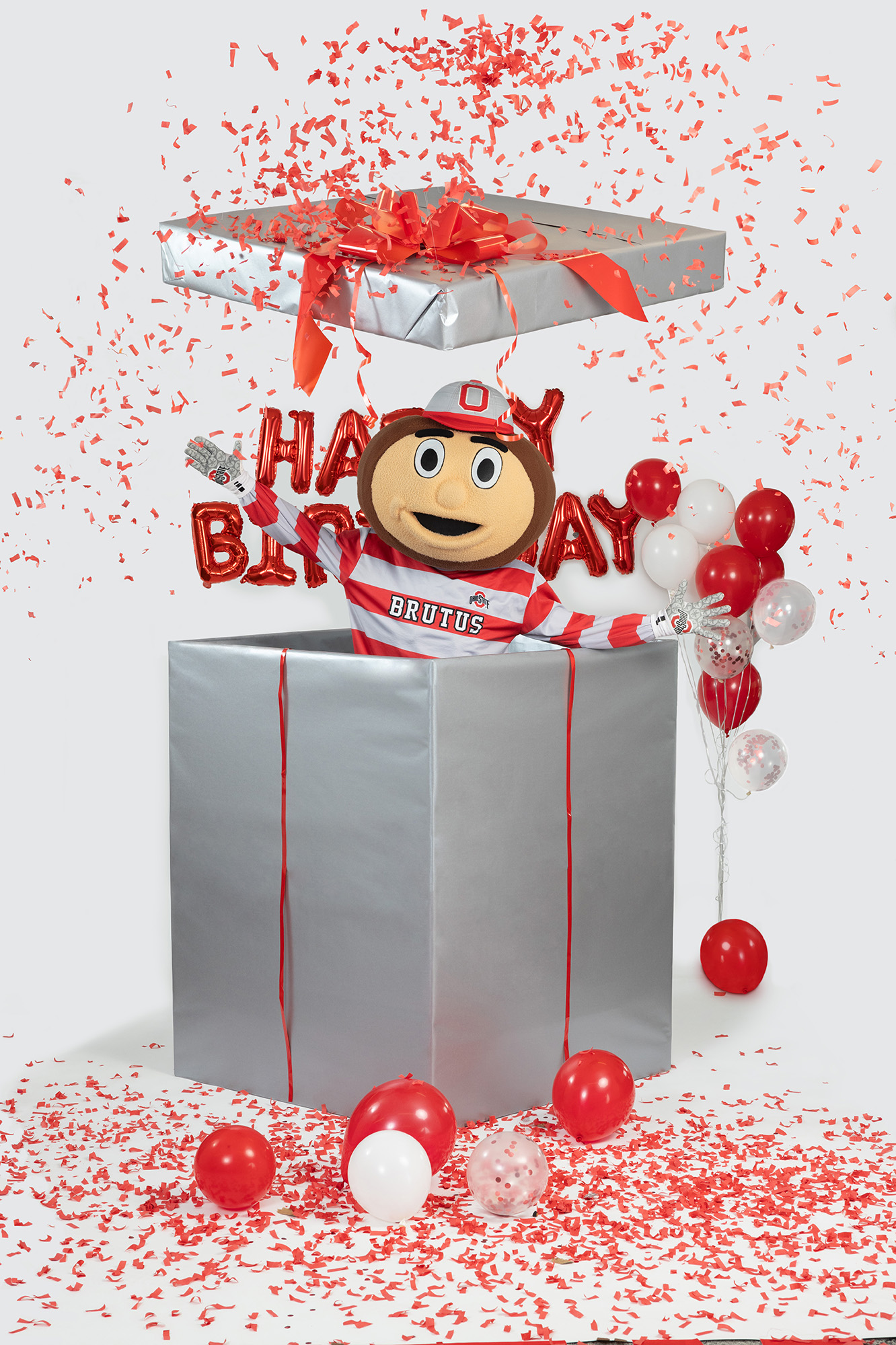 In this Ohio State photo, Brutus Buckeye jumps out of a giant wrapped birthday present. Balloons in Ohio State's colors, as well as shiny mylar balloons that say happy birthday, are around him and confetti flies everywhere. Brutus is the university's mascot--a giant head shaped like a buckeye tree nut with a big grin.