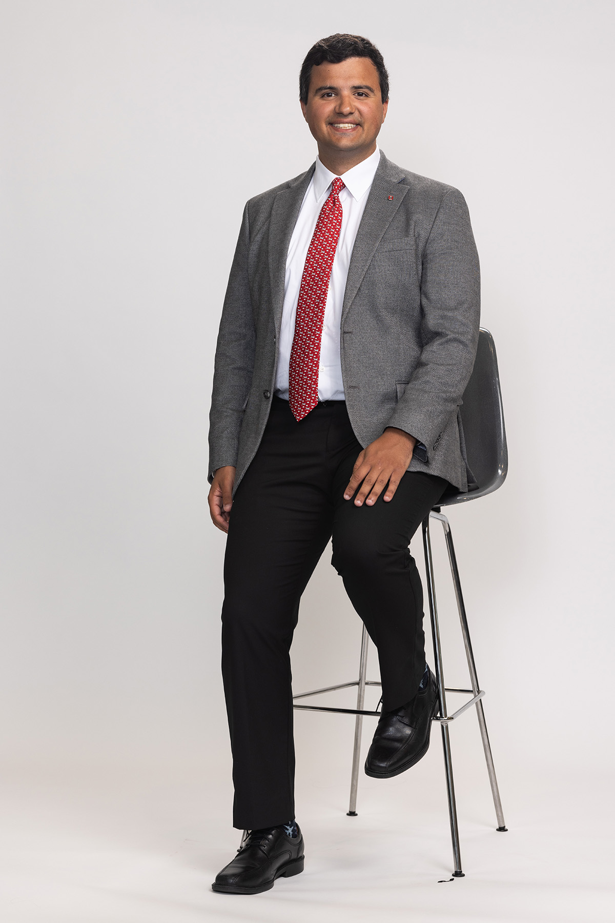 George Valcarel, a young man of Spanish and Greek heritage, sits on/stands against a tall stool. He wears a suit jacket and tie and a smile