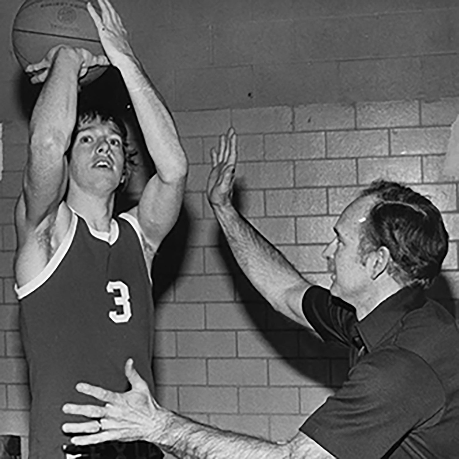 In an older black and white photo, Gene Millard instructs a young man who's holding a basketball as if he's ready to shoot a basket. The coach is either playing defense or correcting the young man's form. He is slightly crouched and has one hand higher in the air. Neither is grinning, but they both look as if they're enjoying themselves.