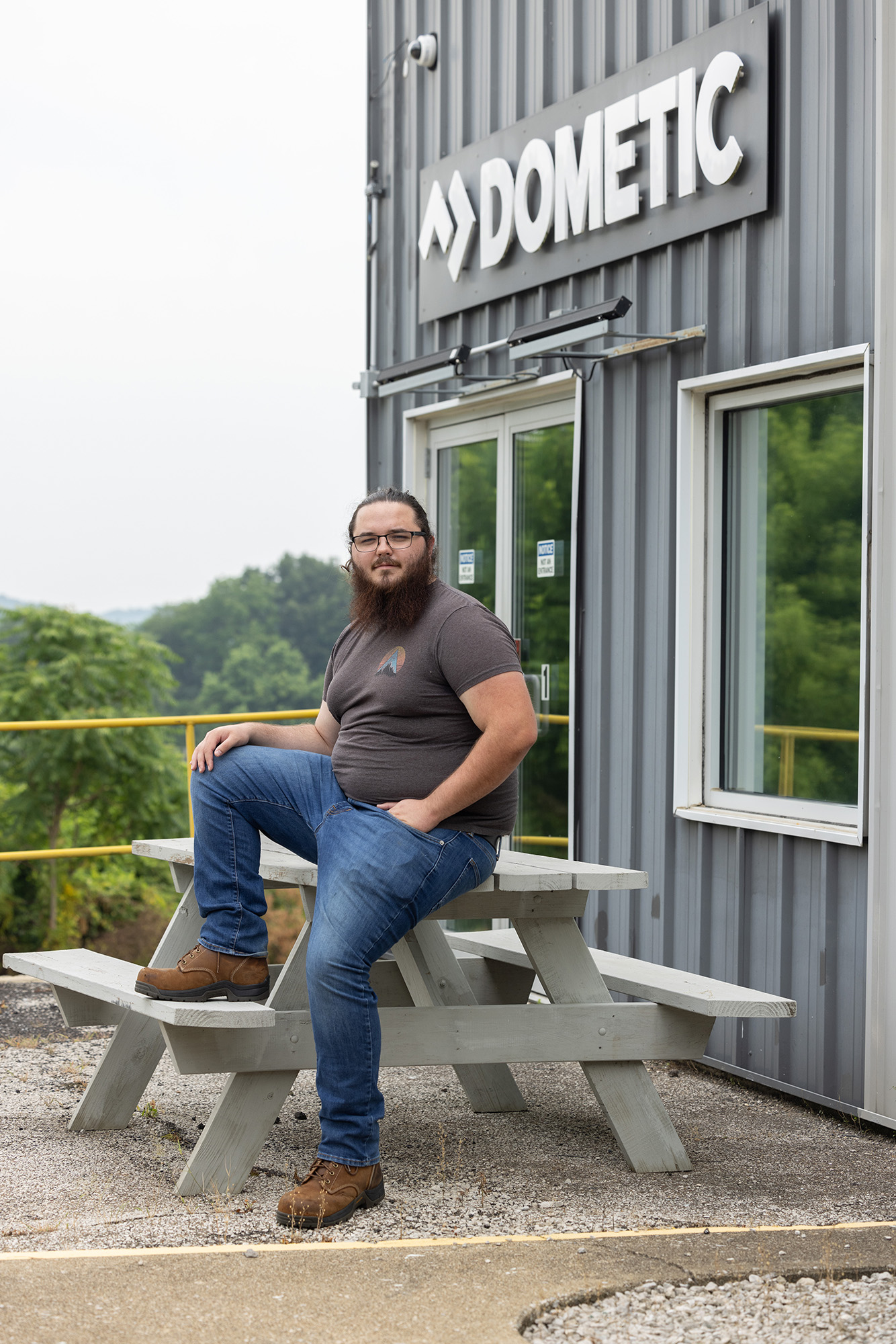 Wearing jeans and a T-shirt, Ethan Montgomery sits on a picnic table outside his work place. He's a white man with long beard and hair pulled back in a ponytail. He has one leg braced on a seat as he sits on top of the table; the other foot is on the ground. He's not smiling but his expression still makes him look capable and friendly.