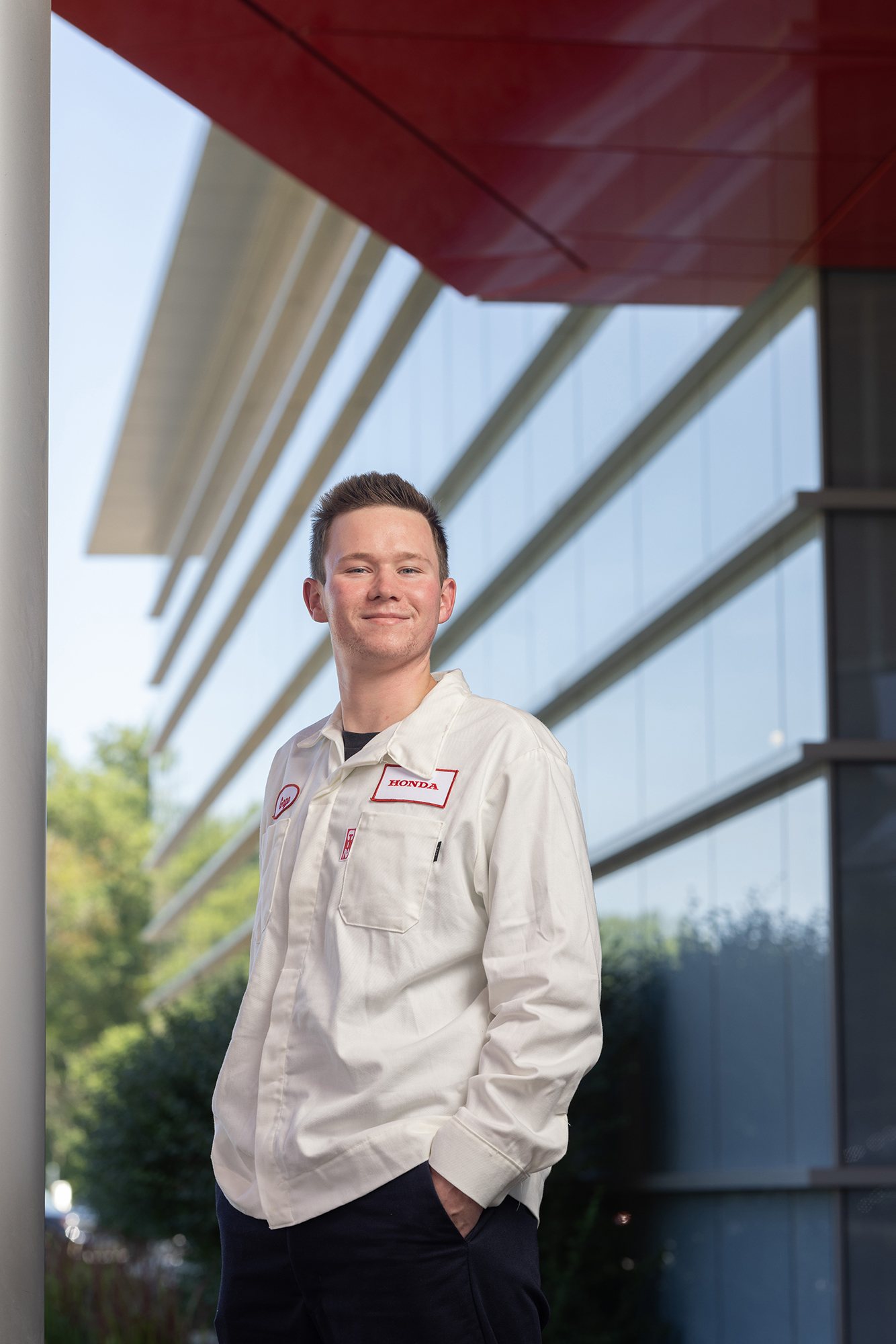 Gage Myers smiles as he looks slightly down at the photographer as he stands outside his workplace. He wears a Honda uniform and has his hands tucked casually in his pockets.