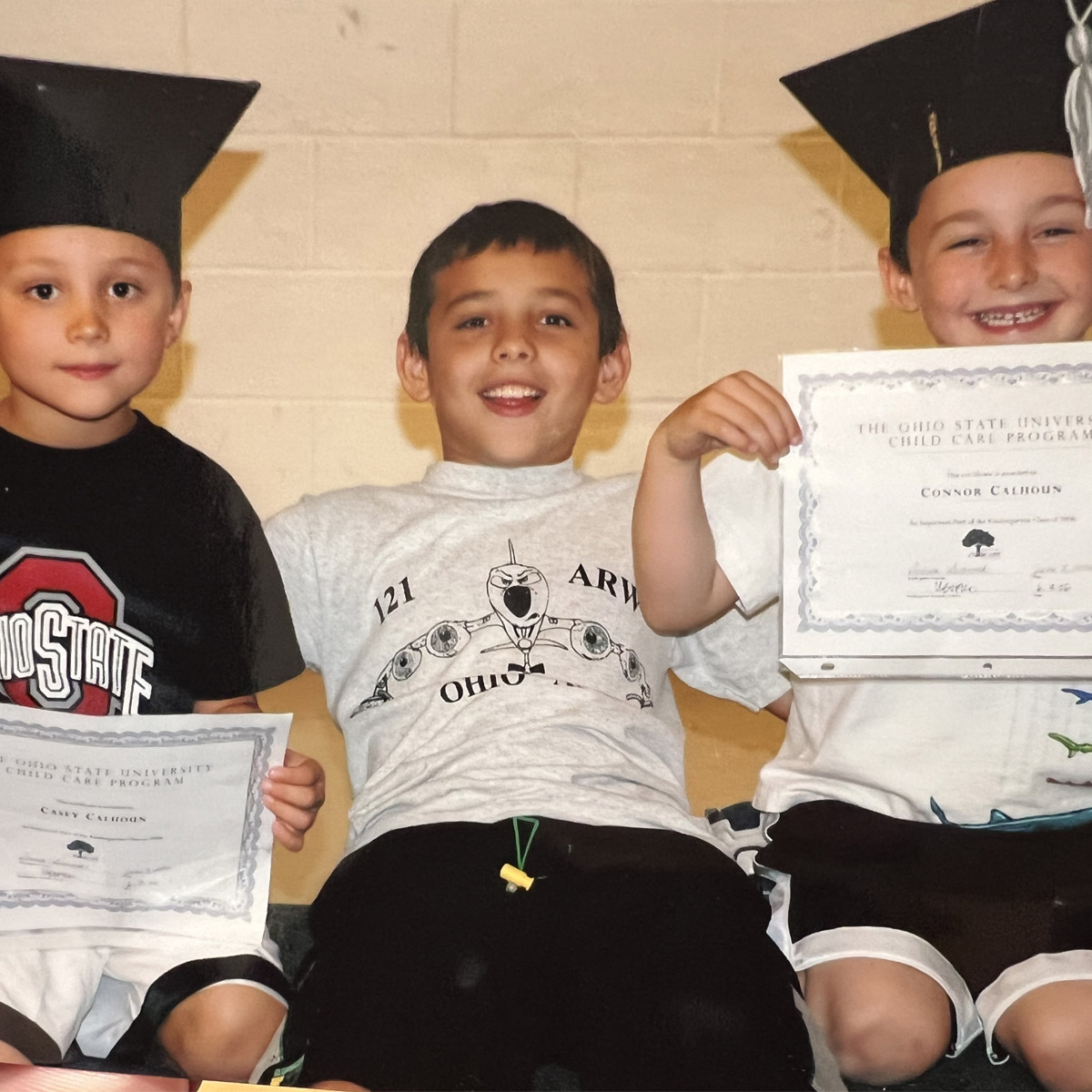 On a bench sit three boys. Two wear graduation caps and show off certificates, and the one between them has his arms wrapped around his brothers’ shoulders. He’s smiling, as is the boy on his left. The expression of the one on the right says he is less enthused, at least at the moment the photo was snapped. 
