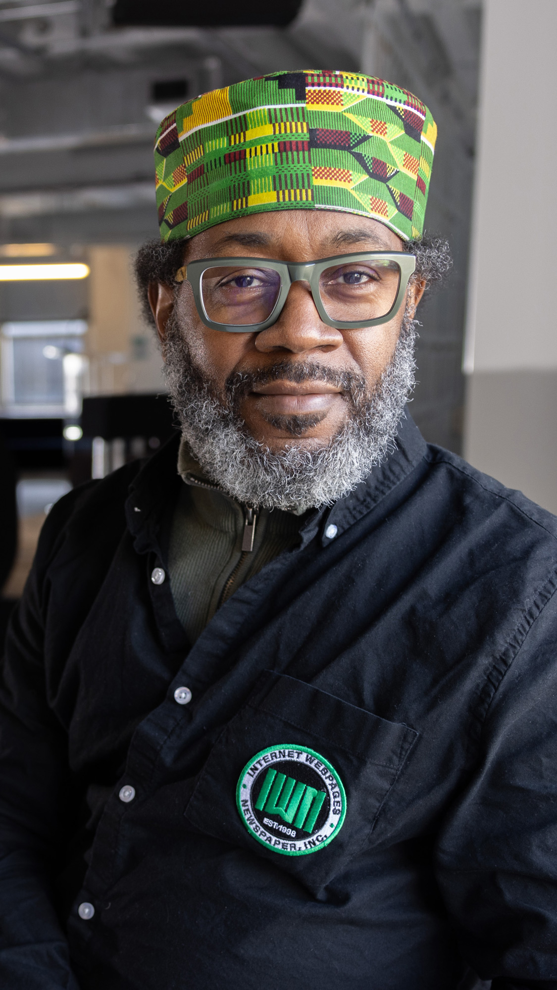 Dante Hamilton, a bearded Black man wearing a kente kufi and thick-frame glasses, poses in an 1871 office area that is mostly in shadow. He has his hands clasped and leans a hip onto a windowsill, so his friendly, quietly confident expression is lit up by sunlight.   