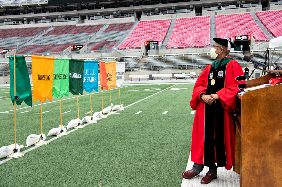 President Drake wearing a black cap and red graduation gown leans against a podium. He is alone in the Ohio State stadium facing a row of flags identifying schools of the university.