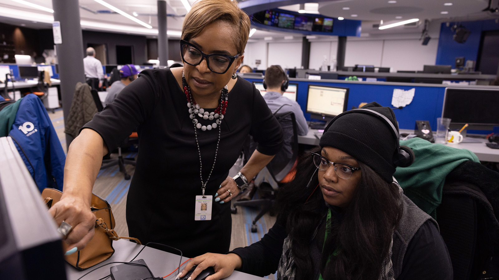 In a brightly lit newsroom, Tracy Townsend points to a computer monitor while working together with another woman sitting at the desk. They’re both wearing glasses and look focused. Tracy is in her on-camera clothes — a black sheath dress and a chunky necklace. The other woman wears a knit cap and vest.
