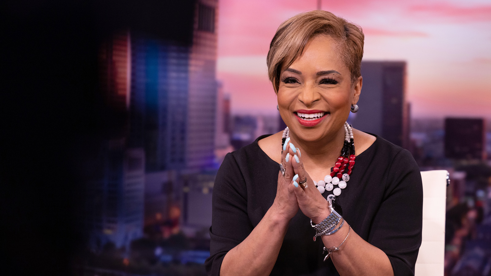 Tracy Townsend, an attractive woman with symmetrical features and bright lipstick, grins as she looks at a camera during the newscast. The background is a picture of a sunrise over Columbus. She touches her hands together in front of her; her long nails are painted baby blue. On the desk in front of her sits a tablet computer.