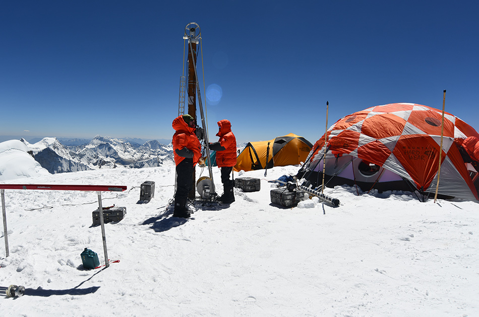 person inspecting an ice drill on a mountain top with tents in the background