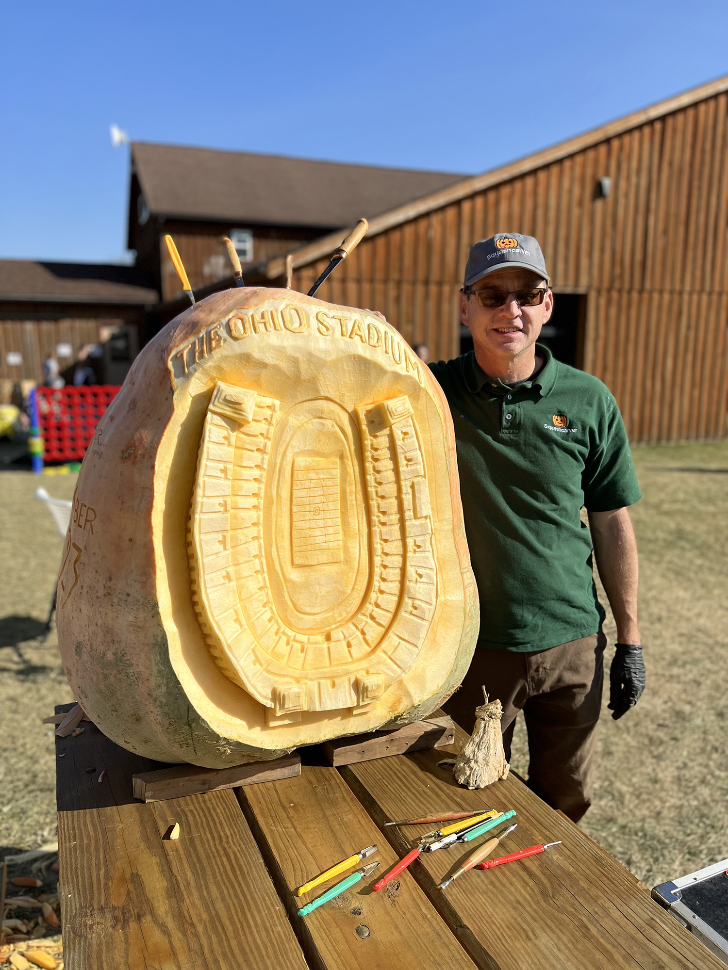 Gus SMithhisler shows off a giant pumpkin he just carved Ohio Stadium onto. He's wearing sunglasses and smiling outside on a very sunny day and his carving tools sit on the picnic table in front of his pumpkin, which is taller than he is. Behind him is a barn.