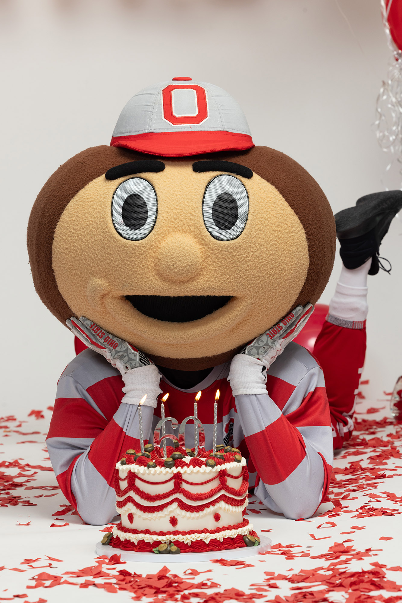 Brutus Buckeye holds his head in his hands as he lies down behind a fancy birthday cake. As always, Brutus is grinning. His pose is similar to a teen girl lying on her bed talking to her best friends--just gleeful.