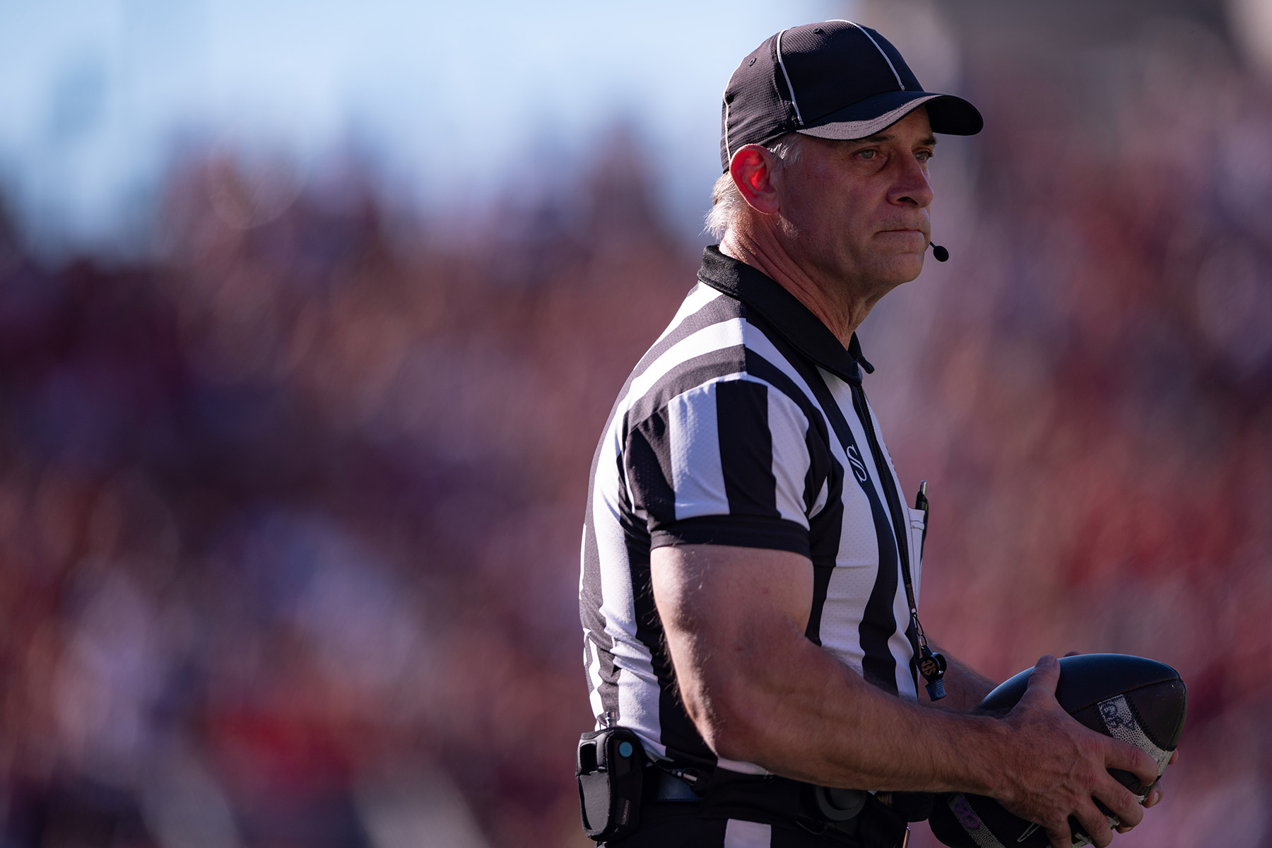 A football referee holds the ball and makes a thoughtful face as he watches something out of the photo frame. He wears a black and white striped referee’s shirt, a ballcap and a headset that leaves a small mic in front of his mouth to communicate calls. He's a white man with white hair and muscular arms.