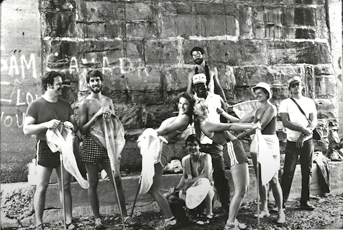 A group of students in swimwear clown around in front of a stone wall along the edge of Lake Erie