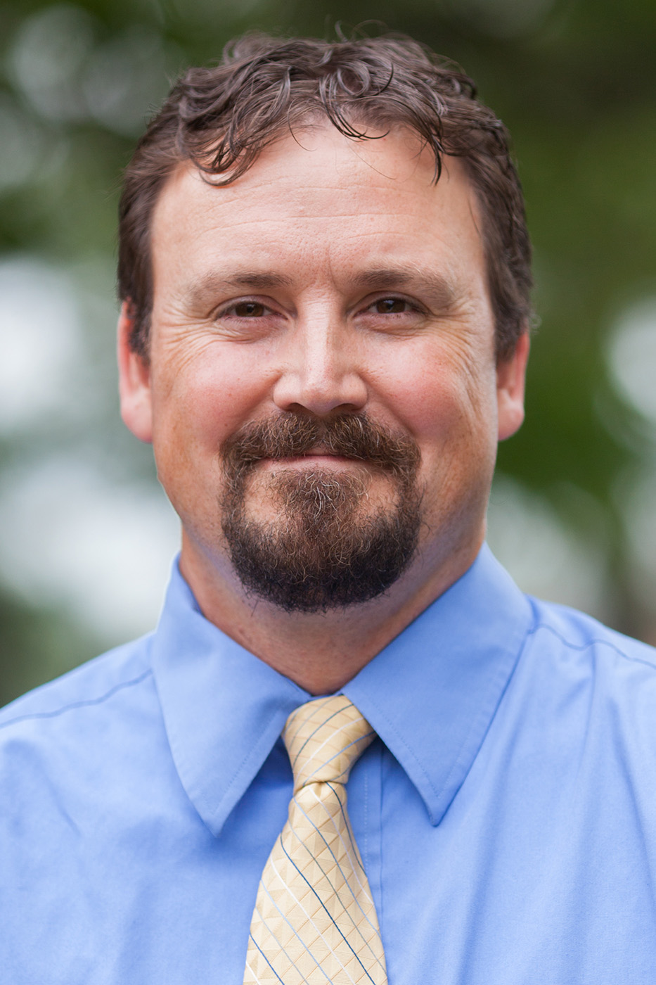 A brown-haired, brown-eyed man smiles for a formal portrait. He wears a shirt and tie.