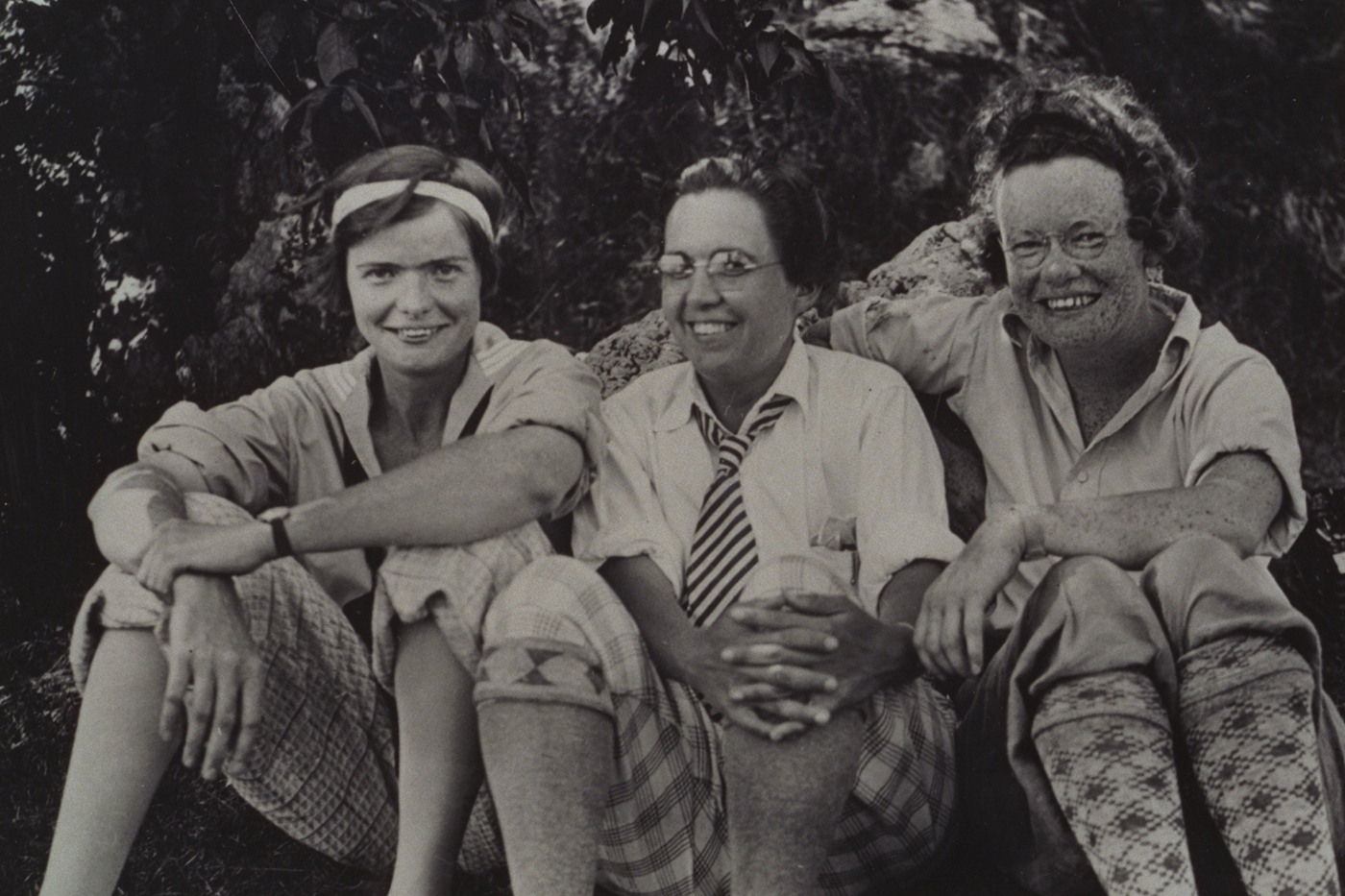 THree smiling white women dressed in 1920s or 1930s clothes sit on a bank