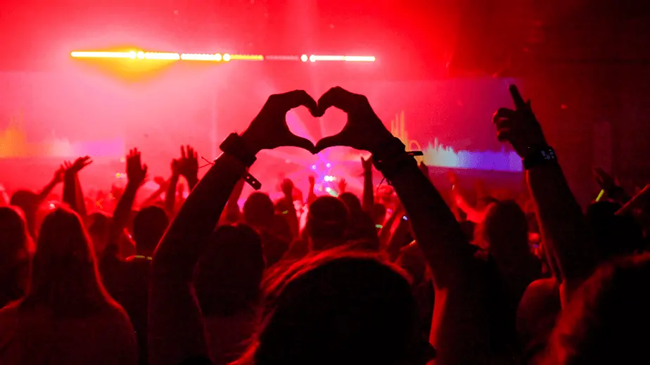 Person making a heart with their hands in a crowd of people dancing by a stage with red lights and fog