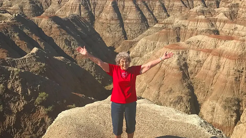 Grandma Joy is standing on top of a canyon peak with her arms raised and outstretched. She is wearing a red shirt and black pants.