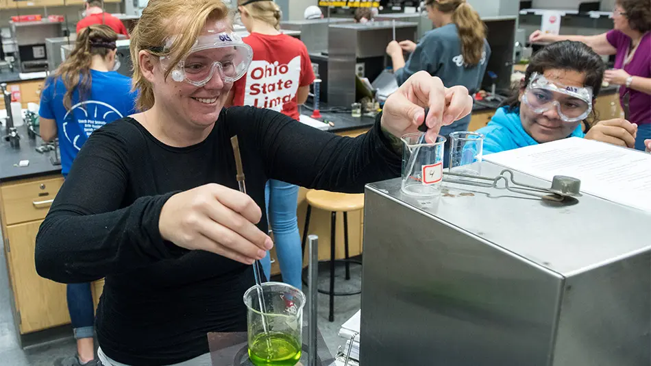 student with protective goggles using glass instrument to stir a green substance in a glass beaker