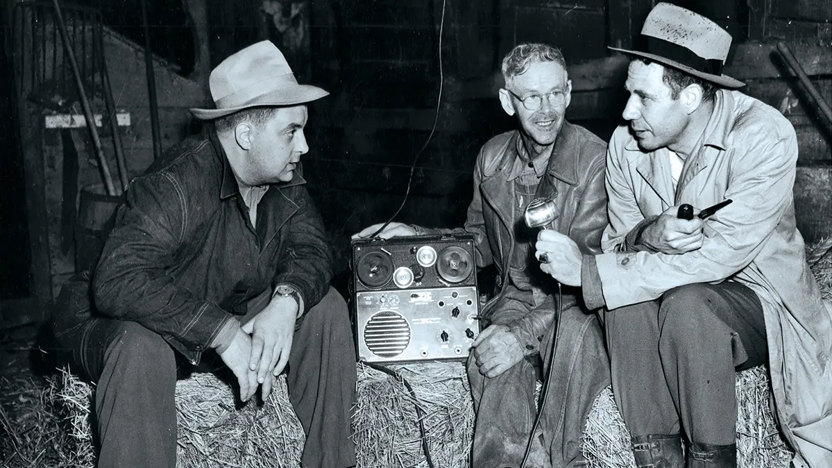 In a black and white photo, three men sit on hay bales in what seems to be a barn. One holds a speaker and another, a microphone. 
