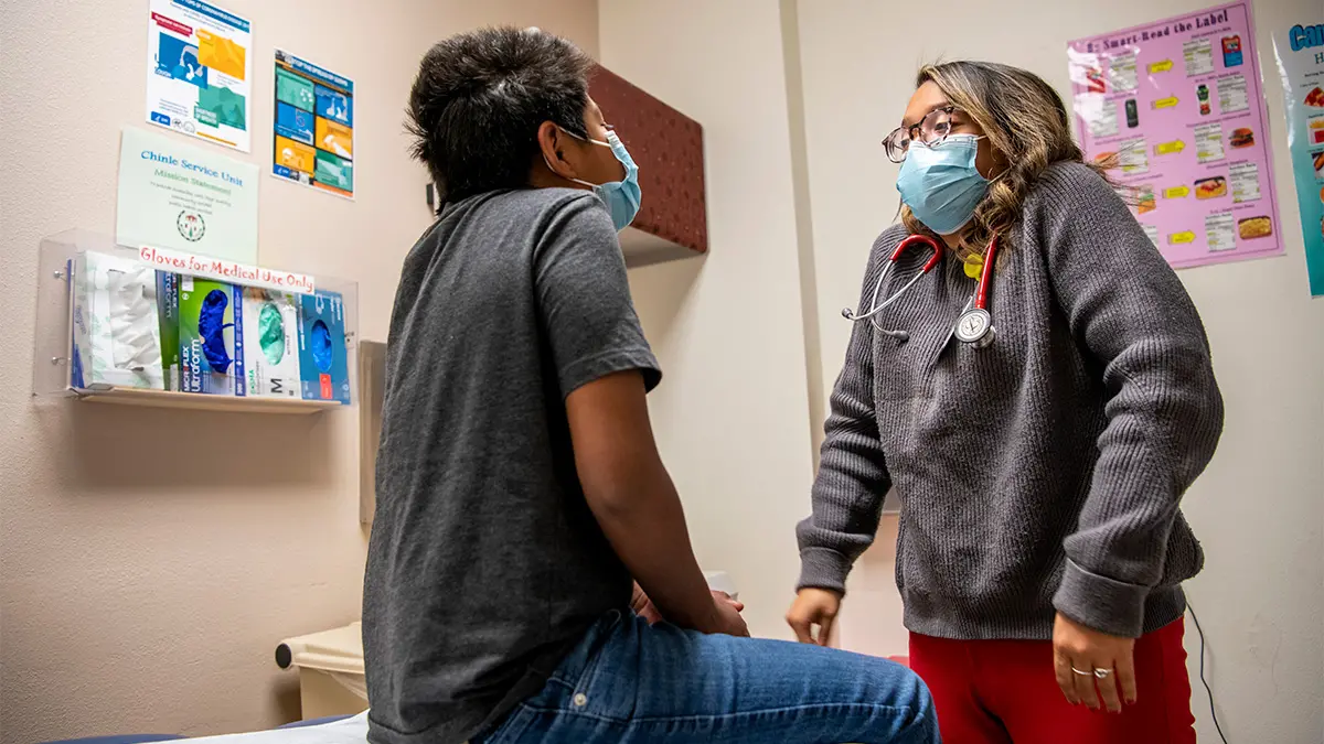 The doctor raises her shoulders to her ears and a male patient copies her during an exam. They’re both wearing gray shirts and surgical masks.