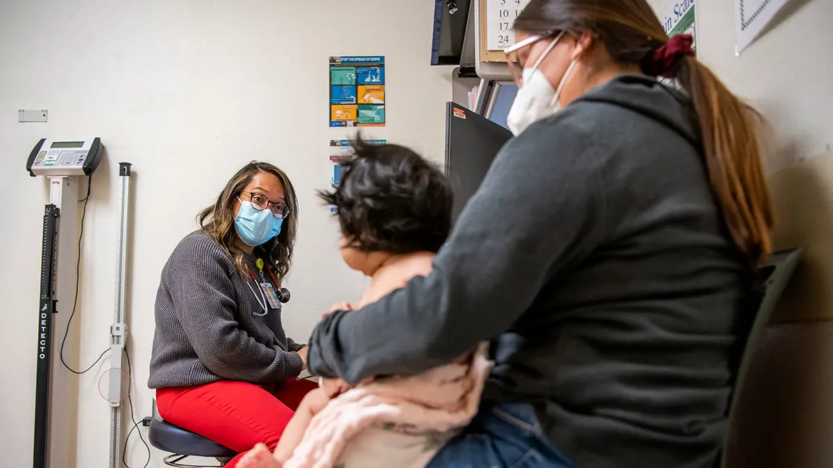 In an exam room, a doctor wearing a mask smiles at a toddler who is sitting on her mother’s lap.
