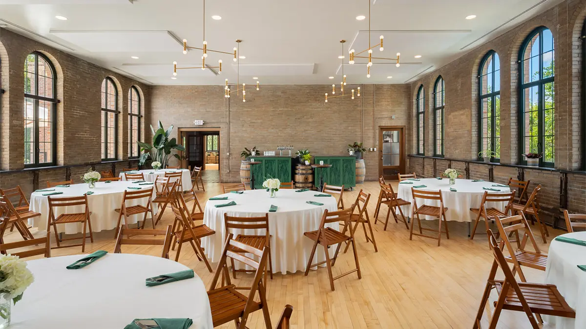 The event space features arched windows, modern chandeliers and tables with floor-length tablecloths and wooden folding chairs. The brick wall and wood floors are muted golds and browns.