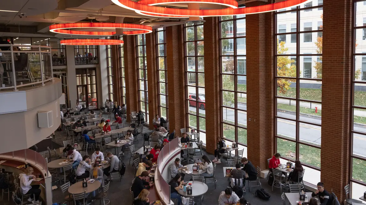 Looking down on a dining room floor from a second-story vantage point, almost three dozen students eat at tables or along counters. Tall windows line the walls and scarlet circles at the ceiling provide more light.