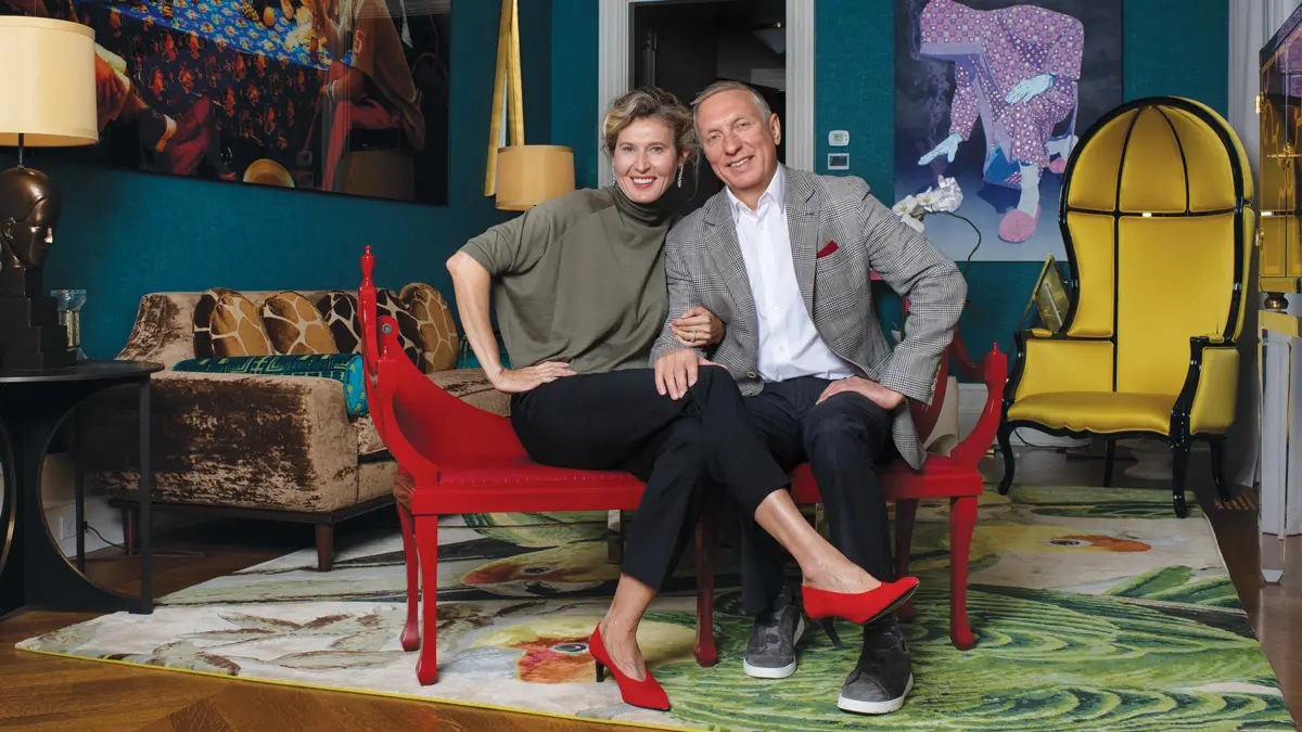 In an elegant room with striking art and bold colors and patterns, a husband and wife sit on a bright red bench smiling. She has her legs crossed toward him and an arm linked with his. His hand is on her knee. Her red lipstick matches her heels and his pocket square.