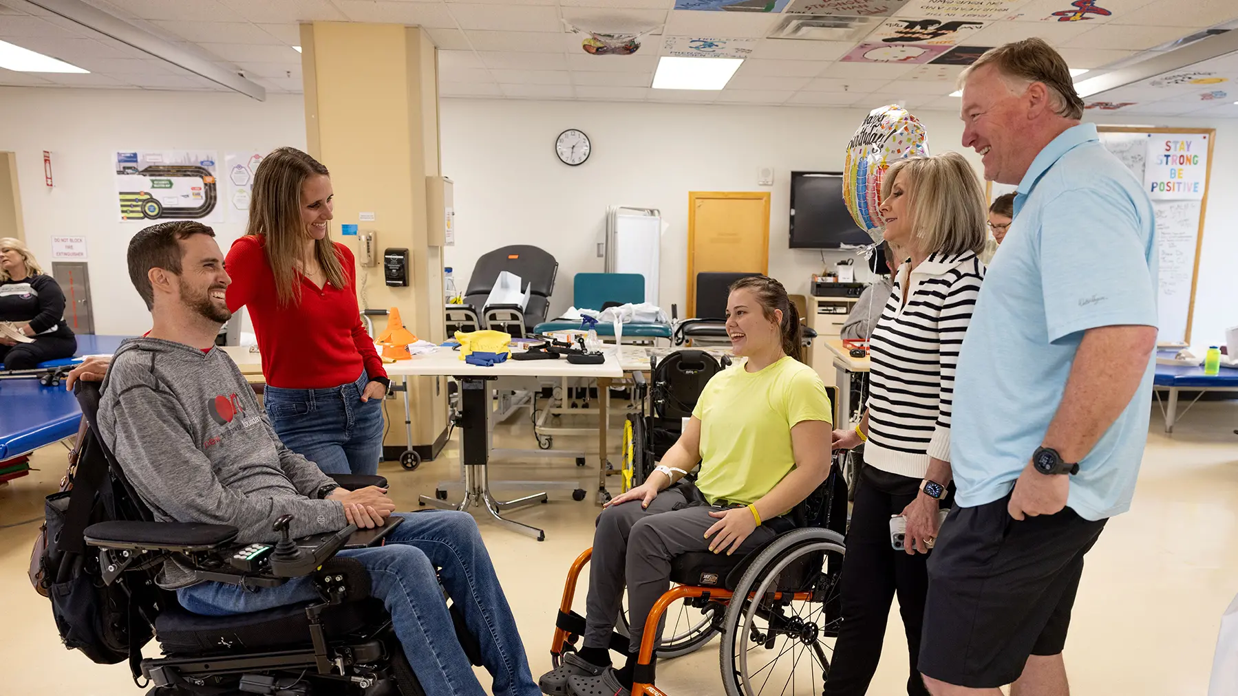 Megan Gentry rests her hand on the back of her husband’s wheelchair as the couple talks with a young woman in a wheelchair, whose parents stand behind her as they all chat. They're in a room with hospital workers and other patients in the background. The three smile and make eye contact as they chat.