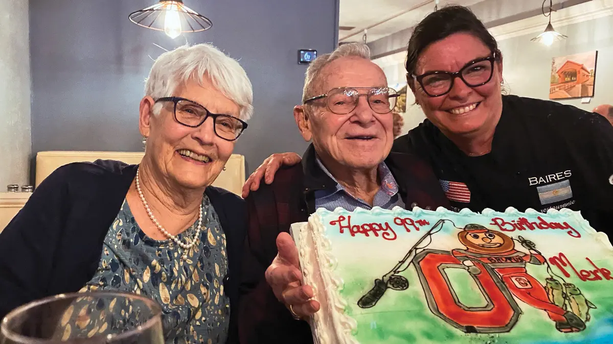 An older couple and dark haired woman smile as they pose for the camera behind a sheet cake. On the cake are Brutus Buckeye, a block O, fishing pole and the words “Happy 99th birthday, Merle.” All three wear glasses and big smiles as they gather close together.