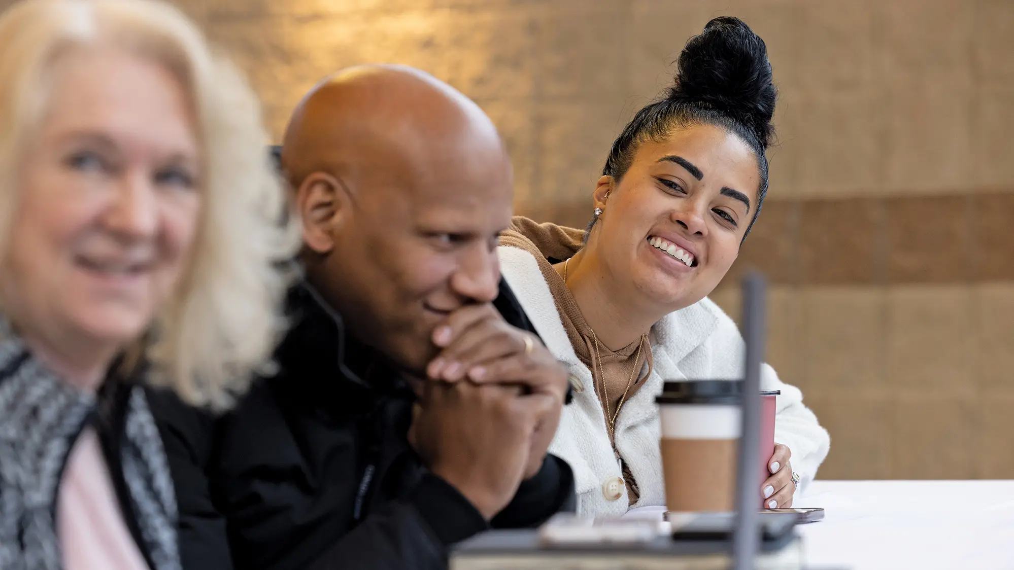 Of the three people in this photo, shown sitting at a table at a meeting, the focus is on a pretty Black woman whose head is wrapped into a high bun on top of her head. She’s smiling like she really enjoys what is being said as beside her, her husband, smiles, too, with his hands folded in front of his mouth. The person closest to the camera is an older blond woman watching the speaker.
