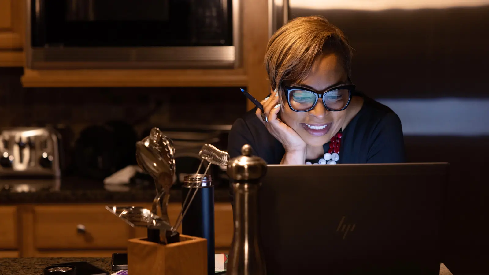 In a dimly lit kitchen, a Black woman with a stylish haircut leans on her counter as she smiles at her laptop, which is showing a virtual meeting with colleagues. The light from the monitor lights up her face and reflects off her glasses.