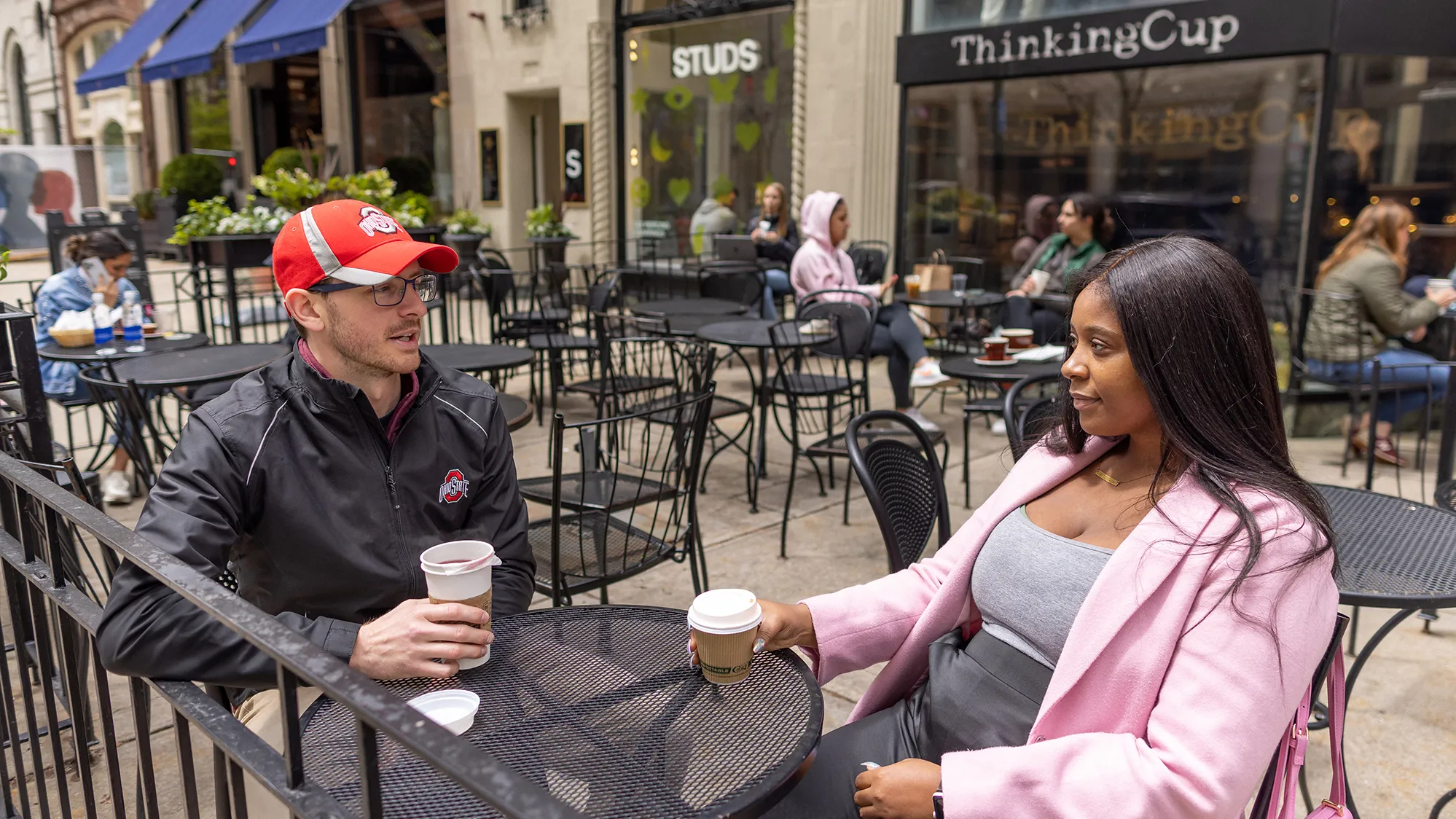 Josh Javor and Dominque McClean talk as they sit a small table in an outdoor dining area on a city block. In the background, the coffee shop sign says, “Thinking Cup.”