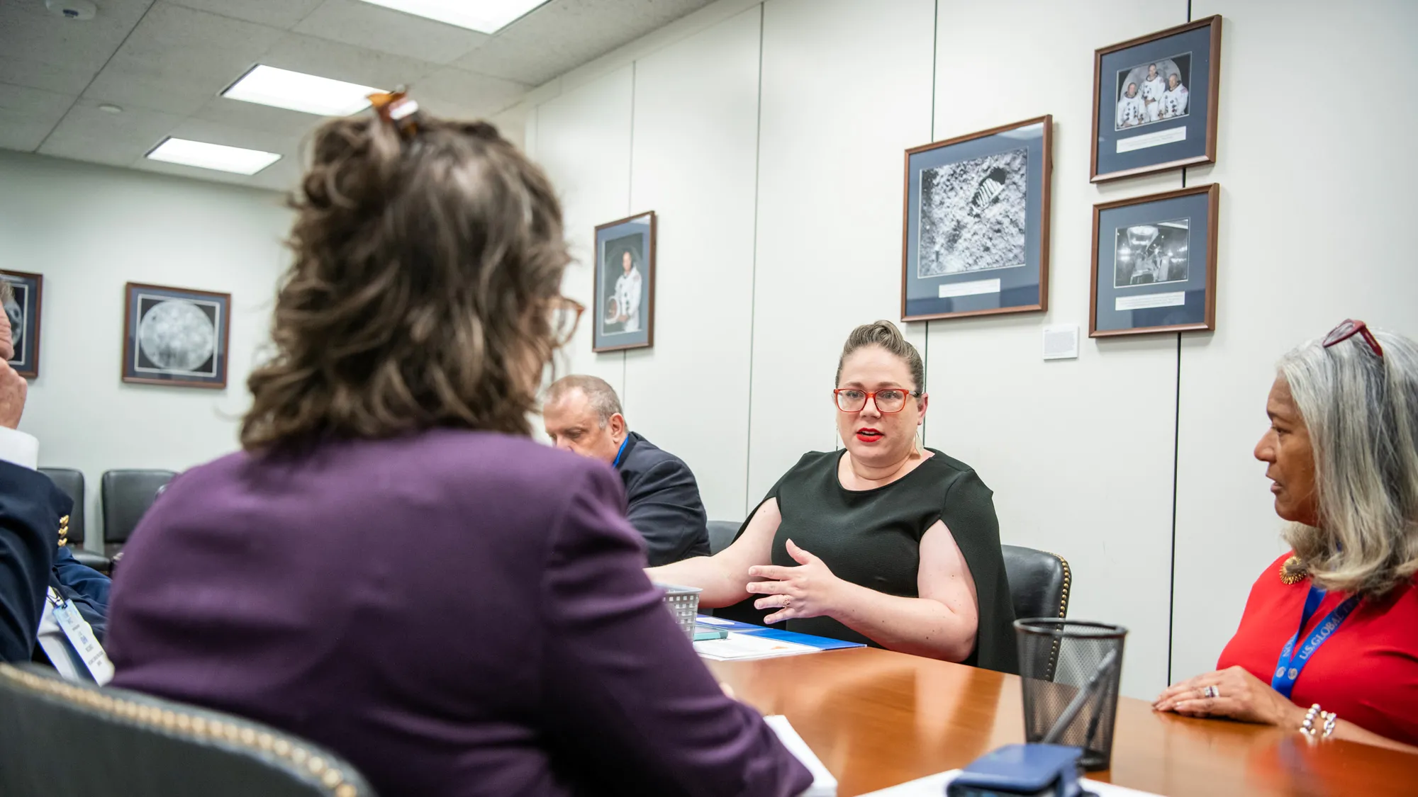 In a small conference room with black and white photos on the wall, Gretchen Klingler sits at a long conference table with other people in business-appropriate clothing. She gestures as she speaks and looks across the table at a listener. 