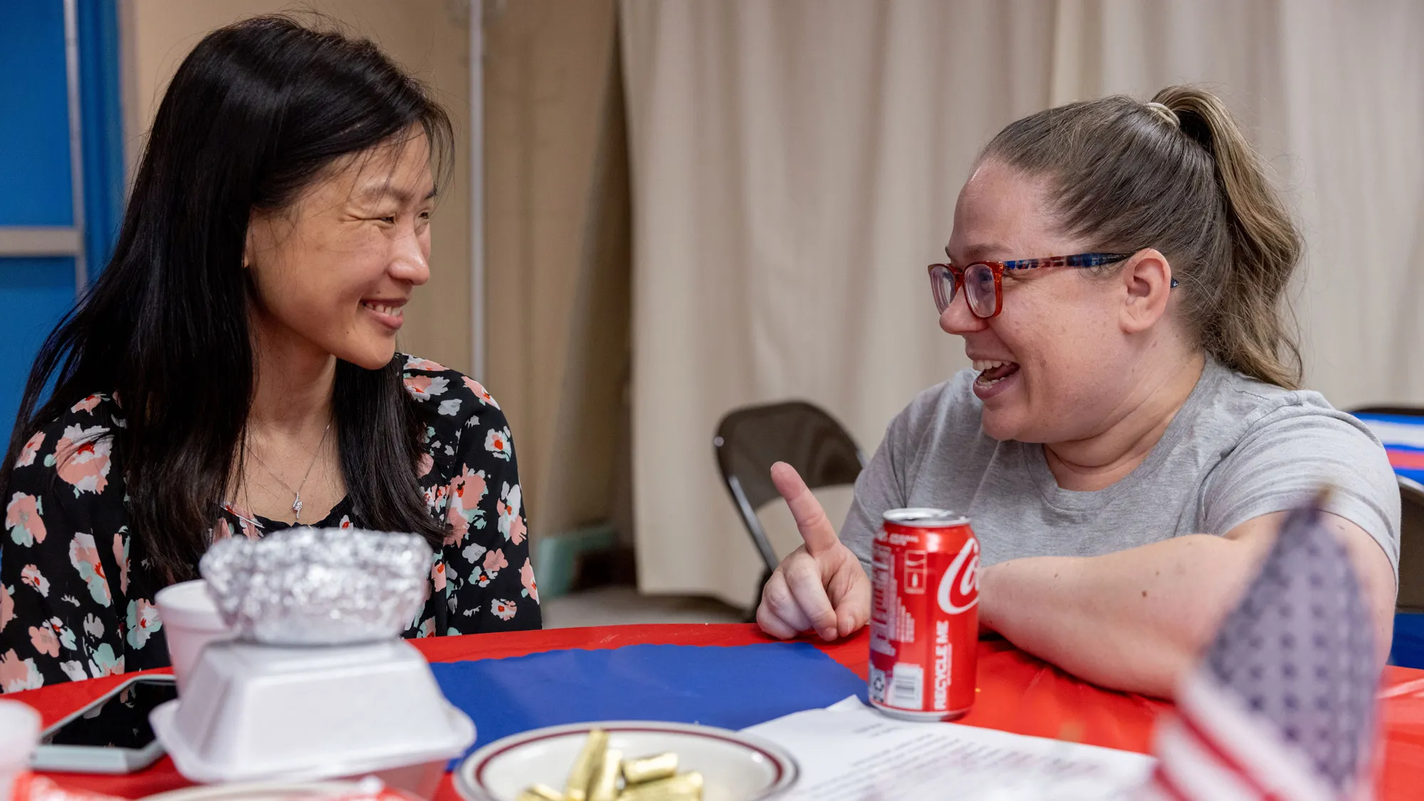 Kneeling next to a table, Gretchen Klingler chats with the woman sitting there. They’re smiling as they focus on each other. The woman is Susan Louderback, a woman of Asian descent with long hair and a flowered top. Gretchen has her hair pulled up in a ponytail. 
