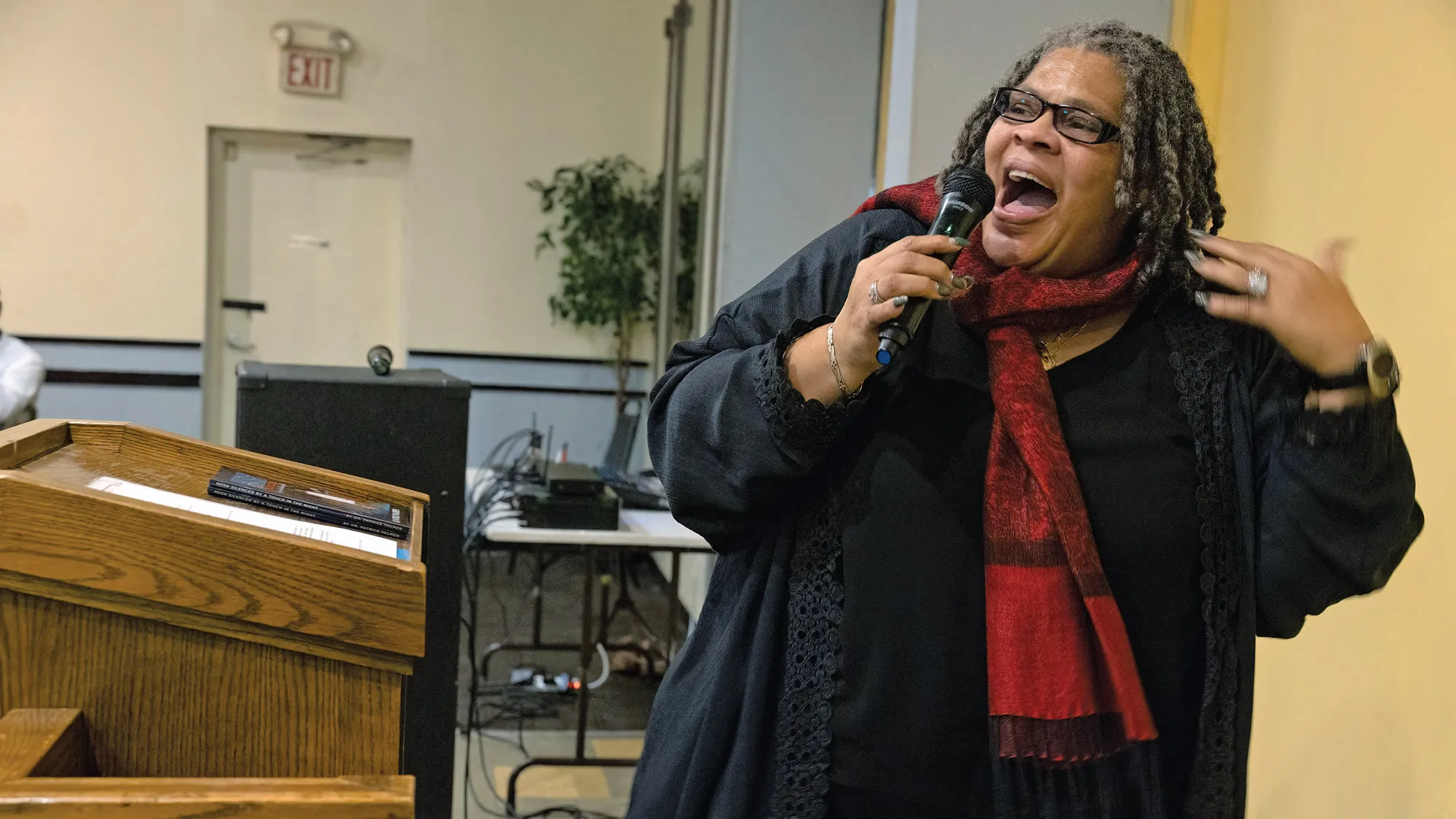 Holding a mic and dressed in flowing clothes with a vibrant scarf, Patrice Palmer gestures as she tells her story. The photograph caught her mid-movement, so she looks like she is stepping forward as she speaks to an audience out of the photo. 