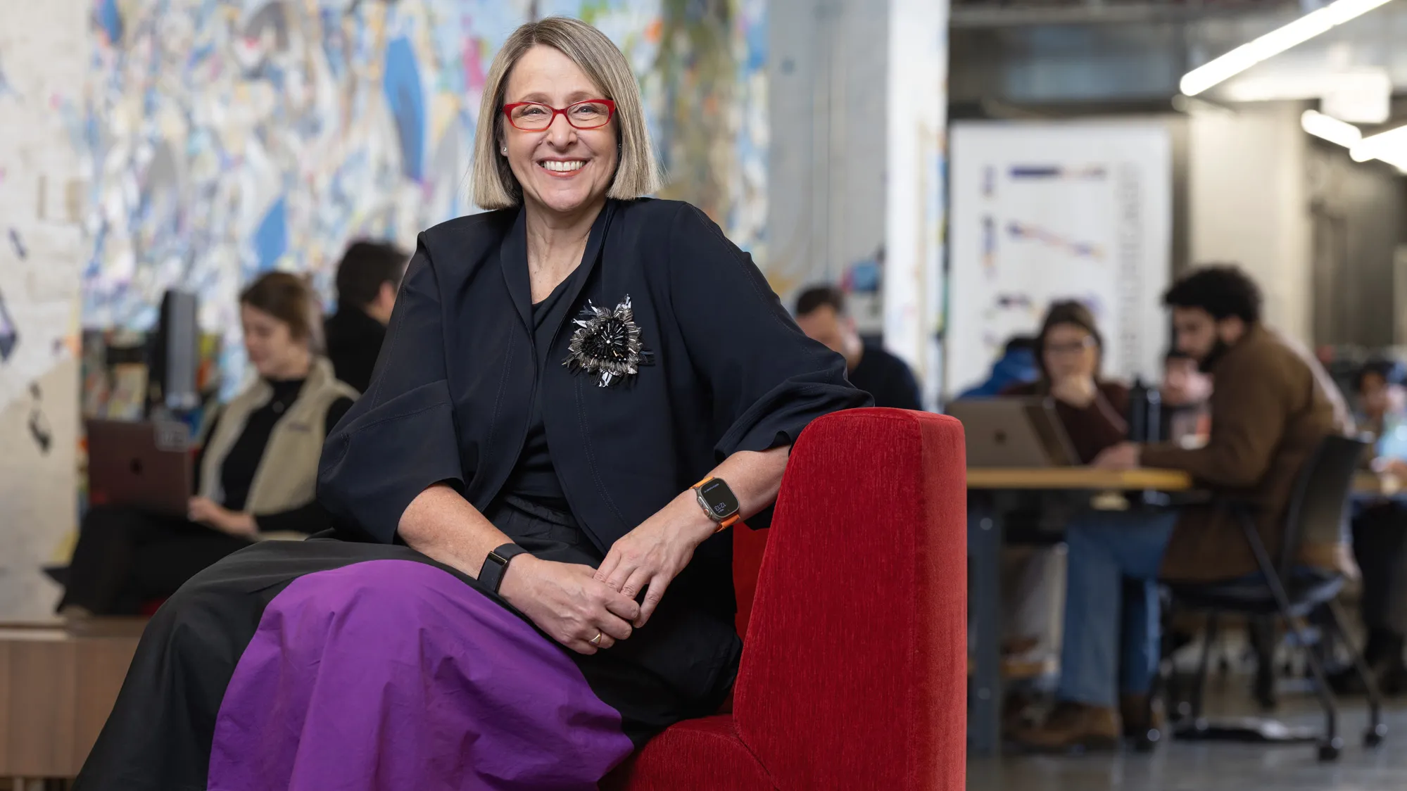 Betsy Ziegler sits on a modern upholstered chair in a space where many people in the background work or chat at small tables. A wall-size painting in the background adds to the lively feel of the scene, while Betsy poses with a genuine smile at the center of it. The middle-age white woman looks comfortable and stylish, with a chin-length haircut, stand-out glasses, modern-shaped clothes and a striking beaded broach that shines but also looks organically shaped.