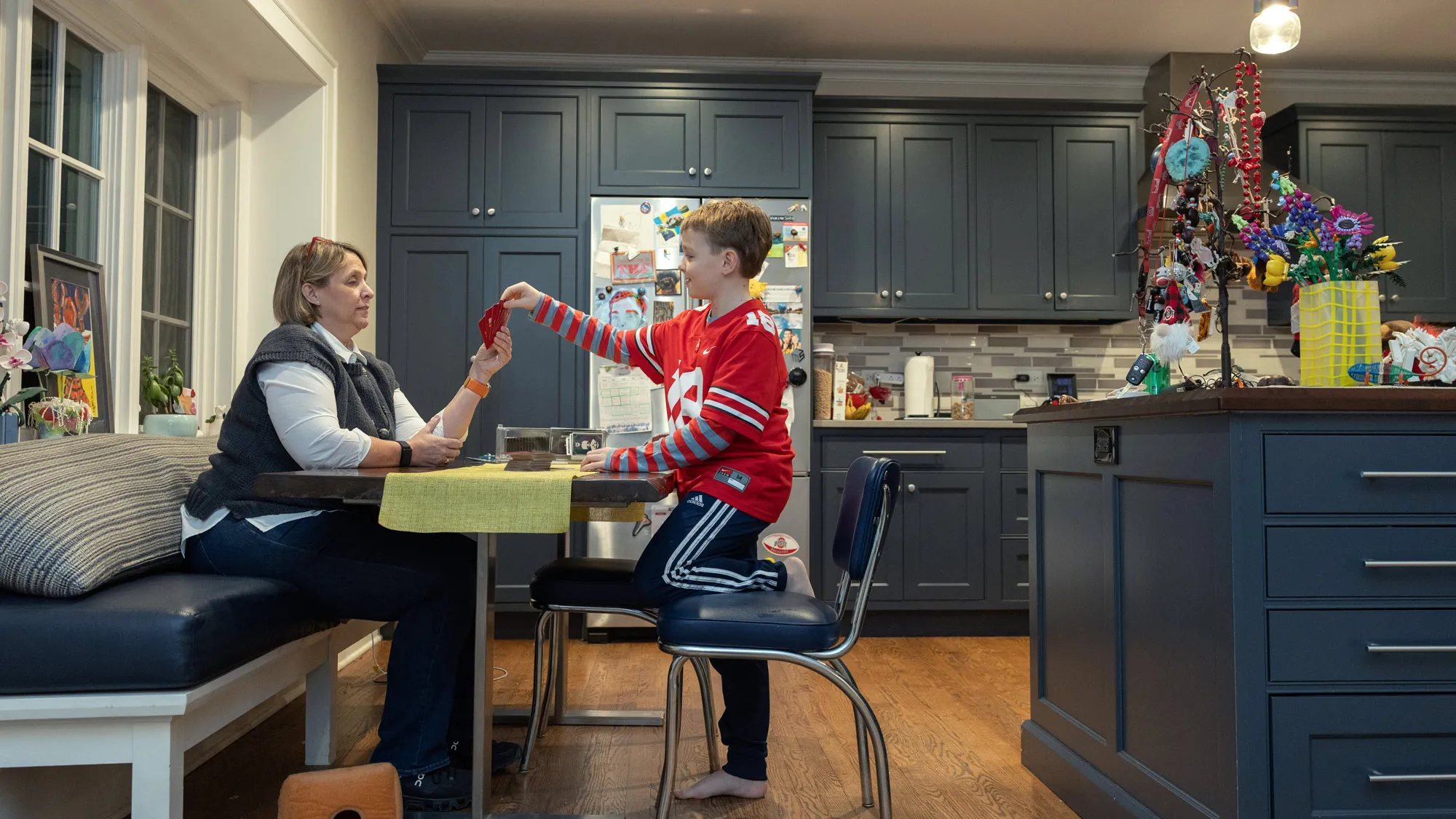 In a beautifully designed kitchen, Betsy sits on a bench at a table holding out a handful of cards to her son, a 9-year-old wearing an Ohio State jersey and sweatpants. They have the same color hair, and seem relaxed, as if this is something they’ve done hundreds of times before.
