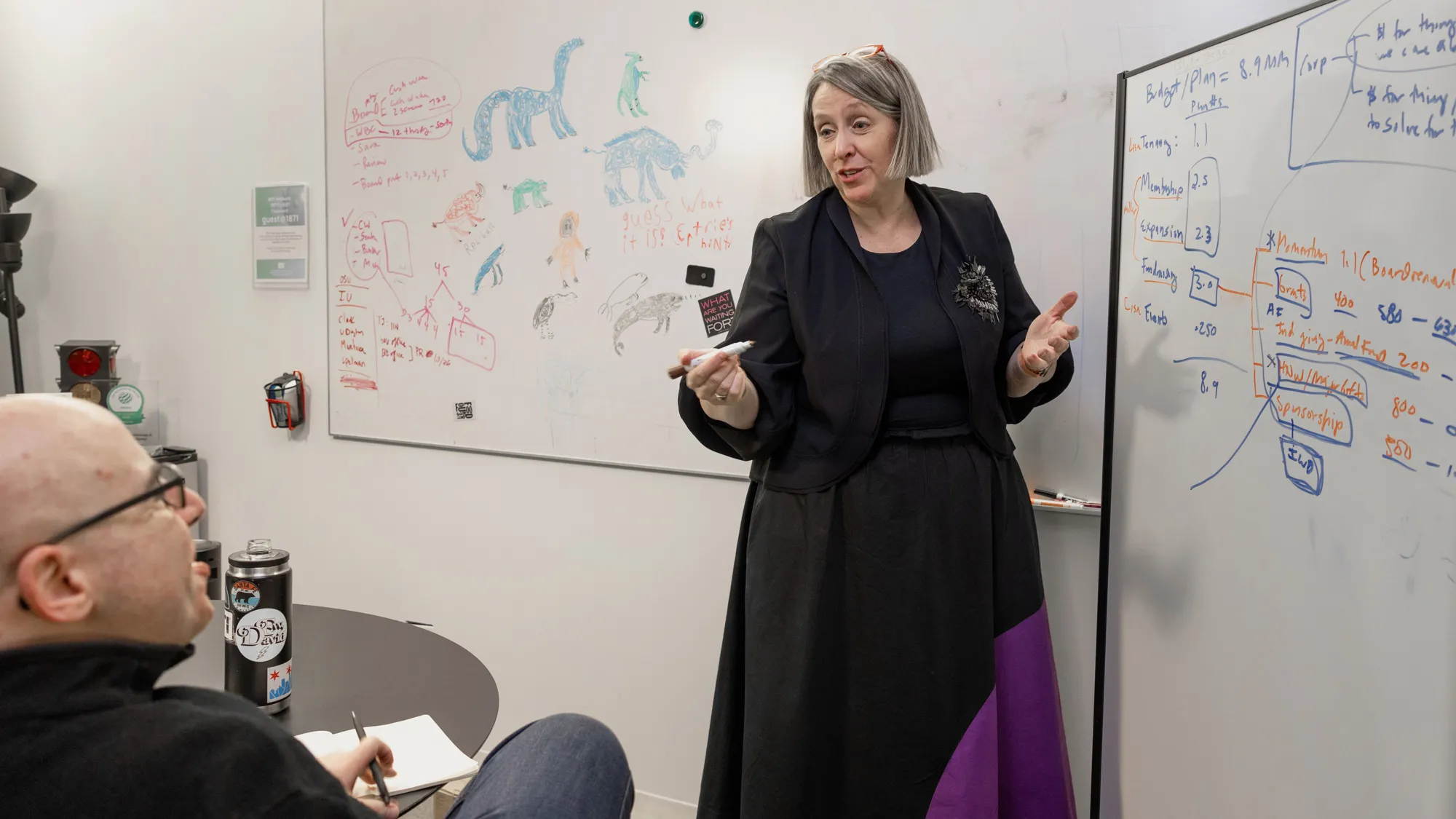 Betsy stands in her office next to a white board, holding an uncapped marker and explaining a point to a man seated at the table in front of her. They’re both smiling as they talk. In the background, another white board has animals and dinosaurs drawn on it (seemingly by a child) next to notes.