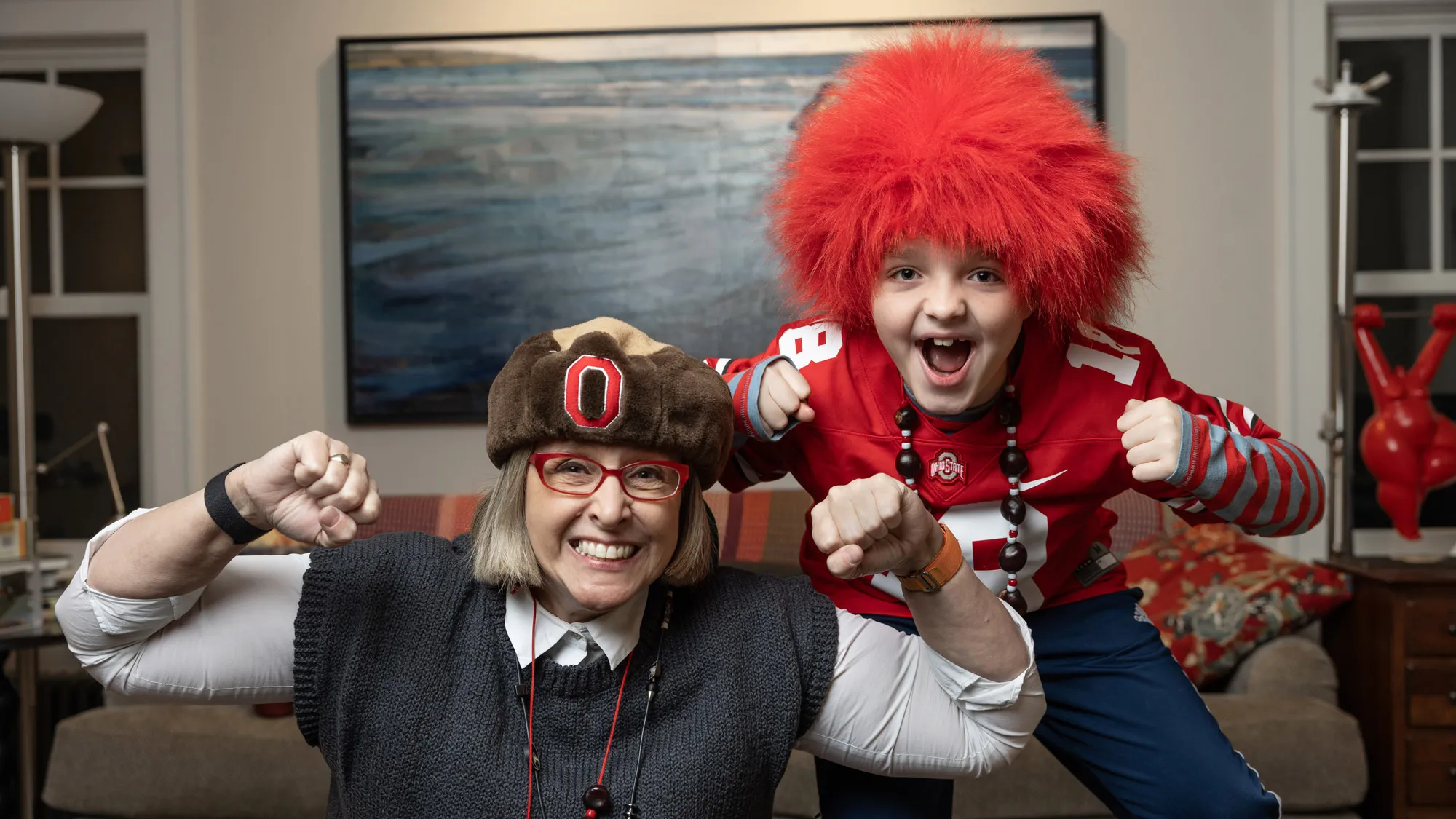 Betsy wears a buckeye necklace buckeye-shaped plush hat and her son Charlie wears a fuzzy, shaggy but short scarlet-colored wig and jersey as they both look into the camera and cheer, as if the camera is a TV showing a game-winning touchdown. They’re in their living room. 