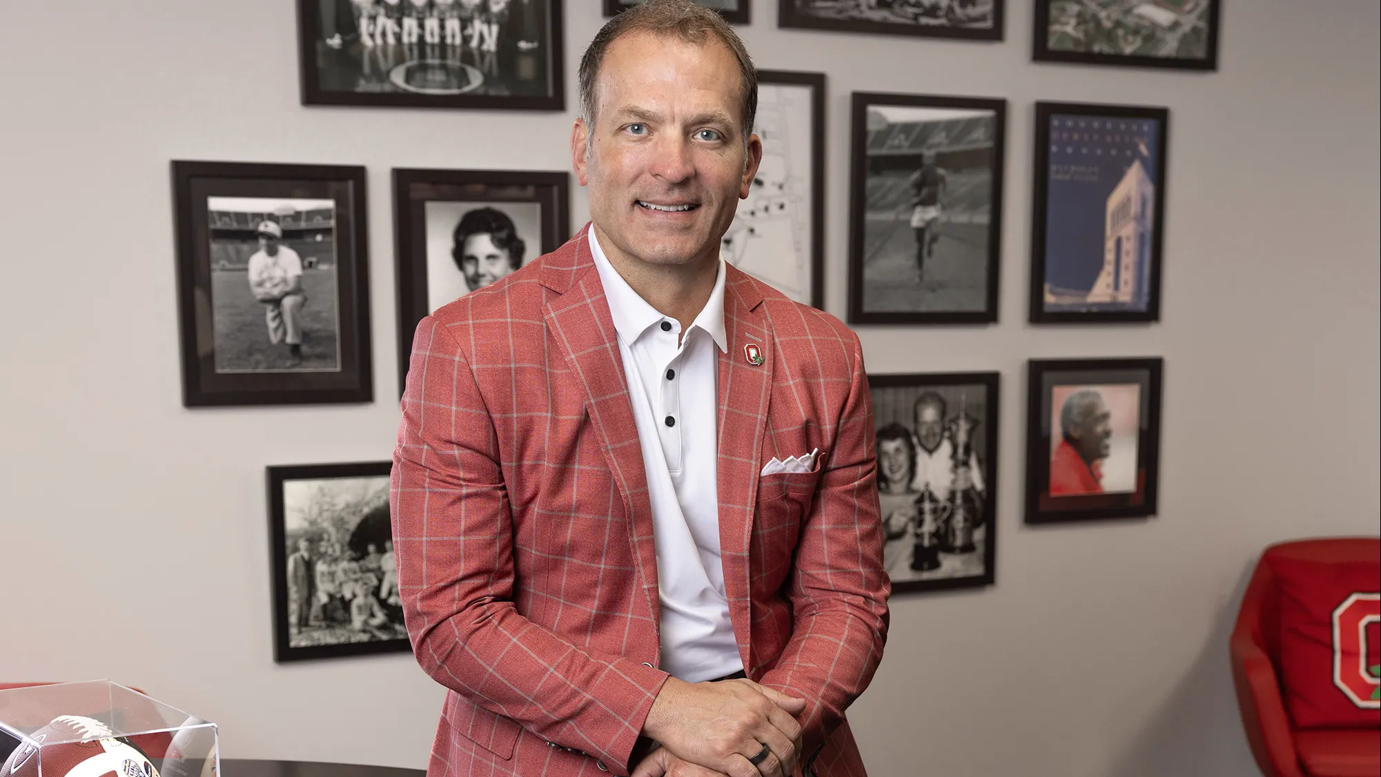 Ohio State Athletics Director Ross Bjork smiles as he poses for a photo in his office. On the wall behind his are photos of great people and moments in Ohio State sports history.