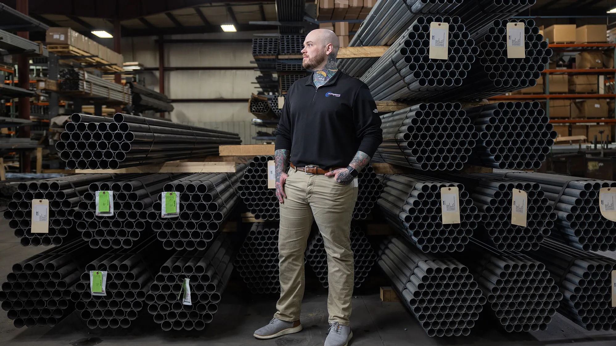 Zachary Ernest, a tall white man with shaved head, neat beard and colorful tattoos on his neck and forearms, is pleased by what he sees on the factory floor of his employer, Ashland Conveyor Products. He stands in front of bundles of about 50 metal pipes each, stacked on shelving.