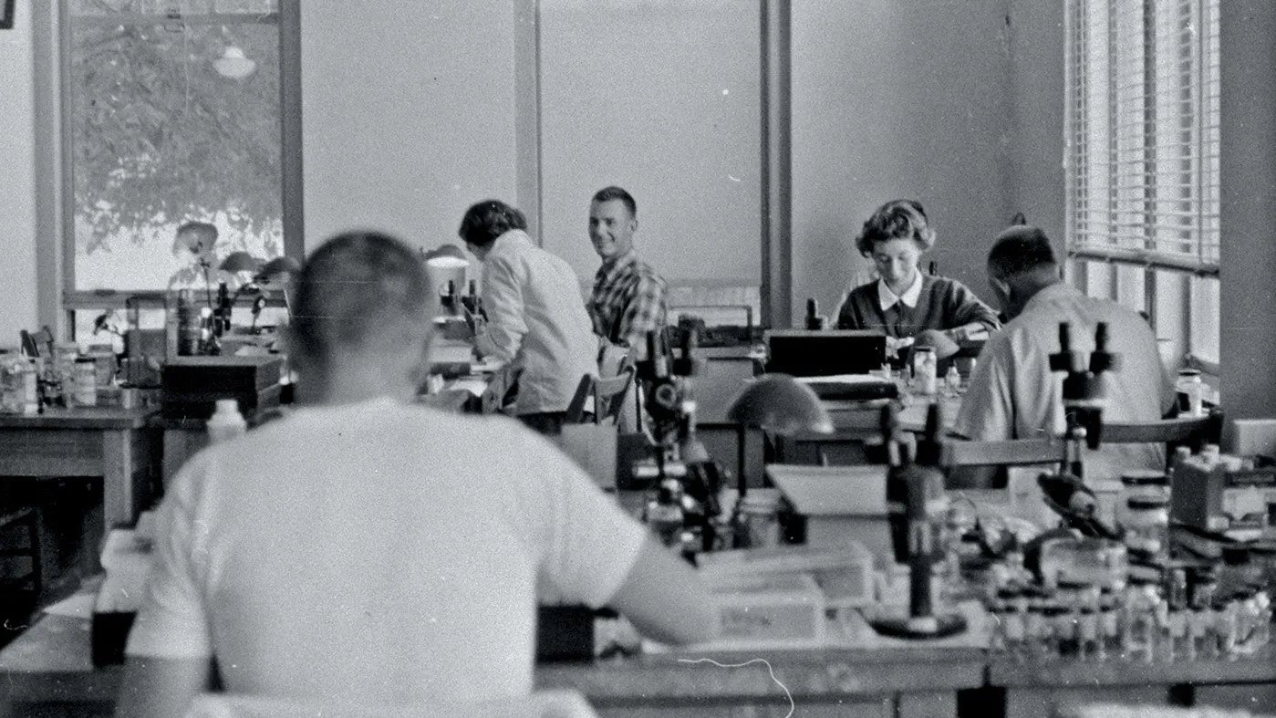 In this old black and white photo from the 1950s or '60s, a handful of students sit at long laboratory tables with all kinds of equipment, including microscopes, spread out.