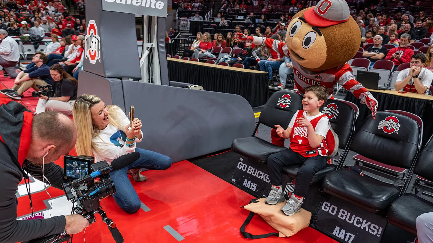 Brutus excitedly poses behind the chair of a grinning young boy who's mid-clap.His mom and a camera man are taing their photo as he sits in special folding chairs set up along the edge of the basketball court