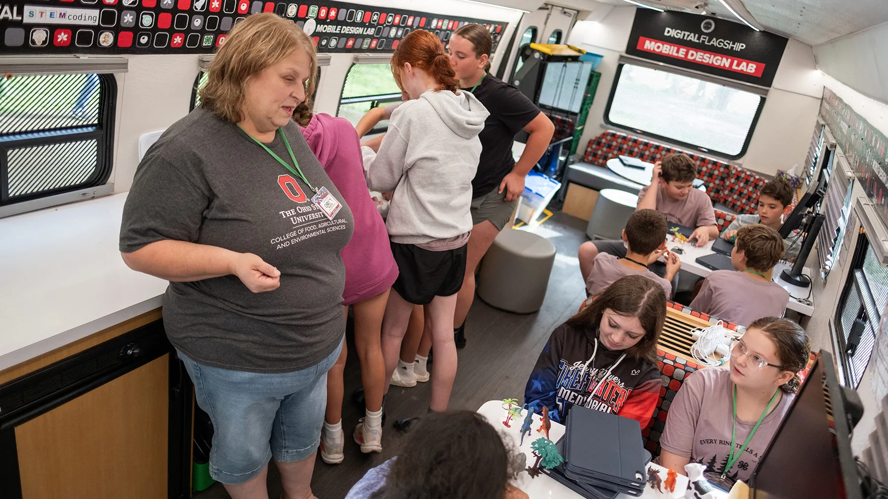 Christy Millhouse smiles as she talks with kids aboard the Ohio State Extension Mobile Design Classroom. It is a big bus filled with tech equipment. About ten kids are using various pieces of the equipment, sitting at tables or leaning against counters.