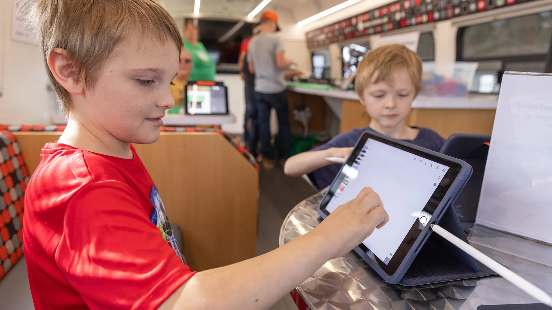 Two boys who look to be brothers use tablet computers aboard the STEM bus. Both look completely absorbed.