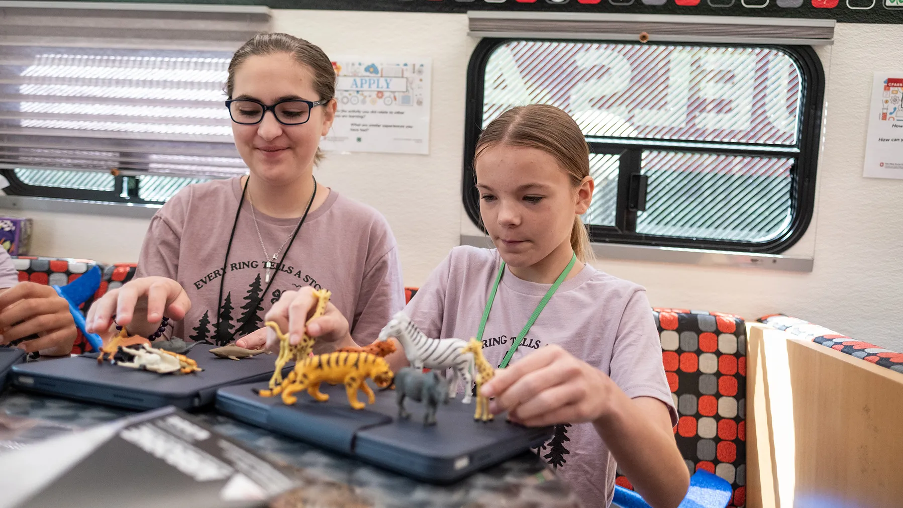 Two girls set up plastic animals on a counter inside the STEM bus, preparing to make a stop-animation movie with tablet computers. They both appear fascinated.