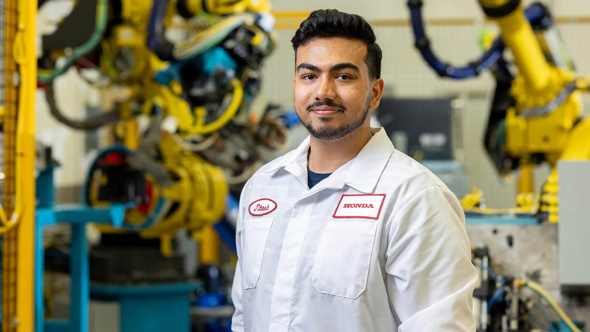 Jitesh Vidhani poses for a photo wearing his Honda uniform and in a hightech factory. Yellow, heavy duty robotic arms and other machinery is working in the background. Jitesh is slightly smiling, with his eyes and mouth.