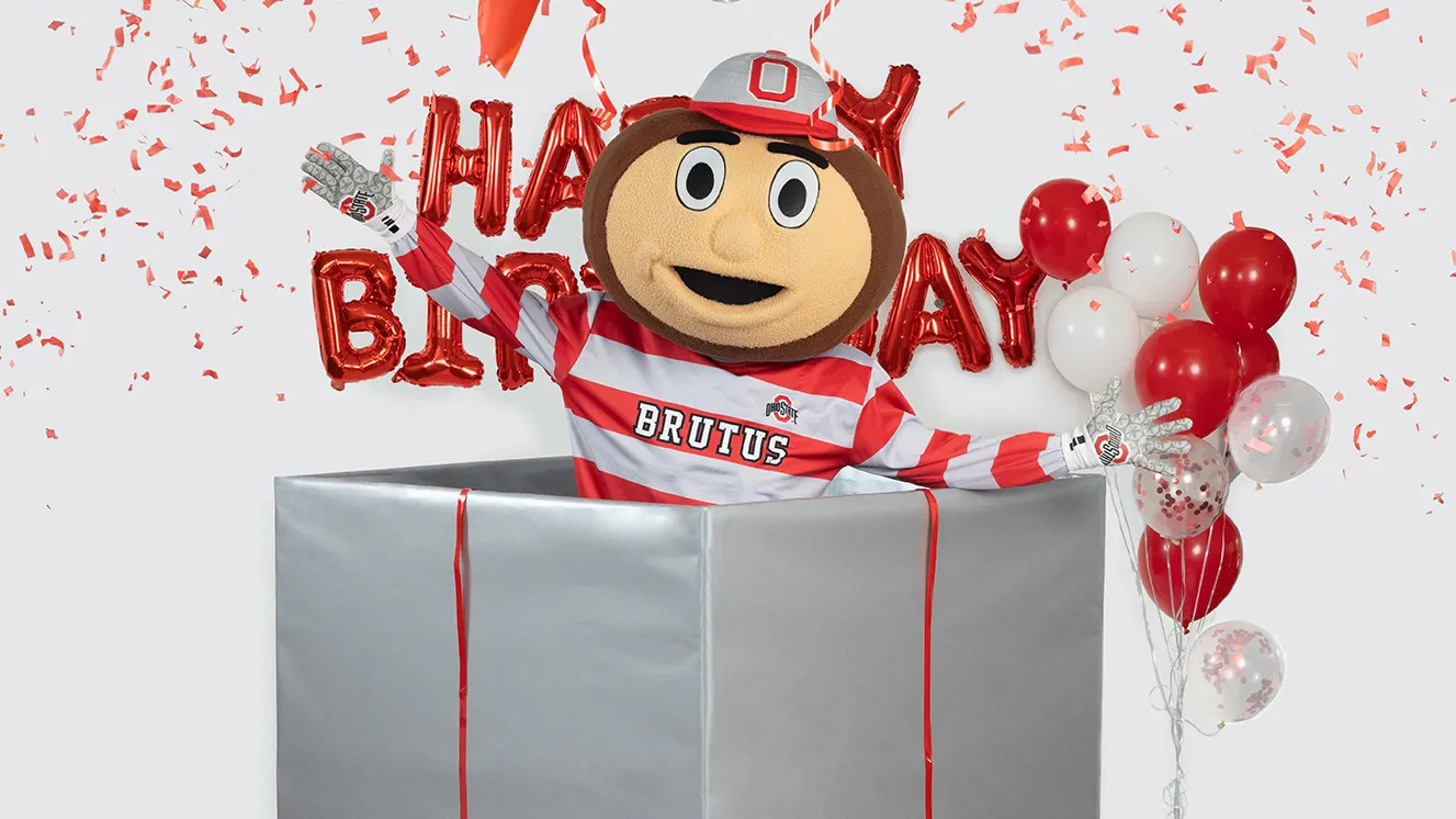 In this Ohio State photo, Brutus Buckeye jumps out of a giant wrapped birthday present. Balloons in Ohio State's colors, as well as shiny mylar balloons that say happy birthday, are around him and confetti flies everywhere. Brutus is the university's mascot--a giant head shaped like a buckeye tree nut with a big grin.