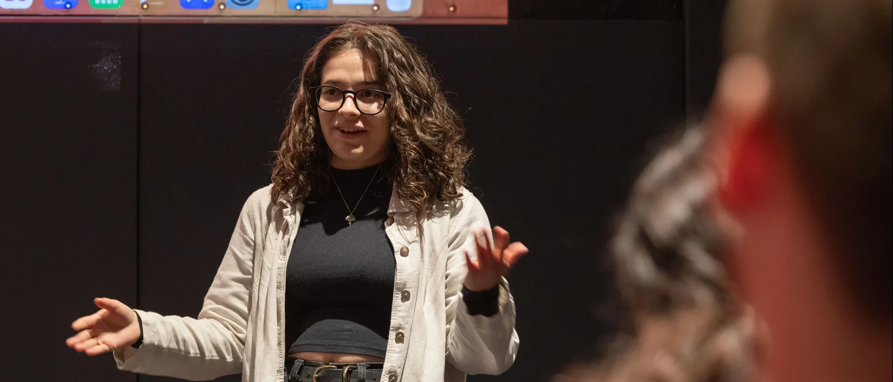 A young woman explains an element of the personal documentary she created to her class in the new building’s screening room. The audience is just hinted at in this photo as the photo is take from behind some students. Cleo, the main focus, gestures and smiles as she talks.