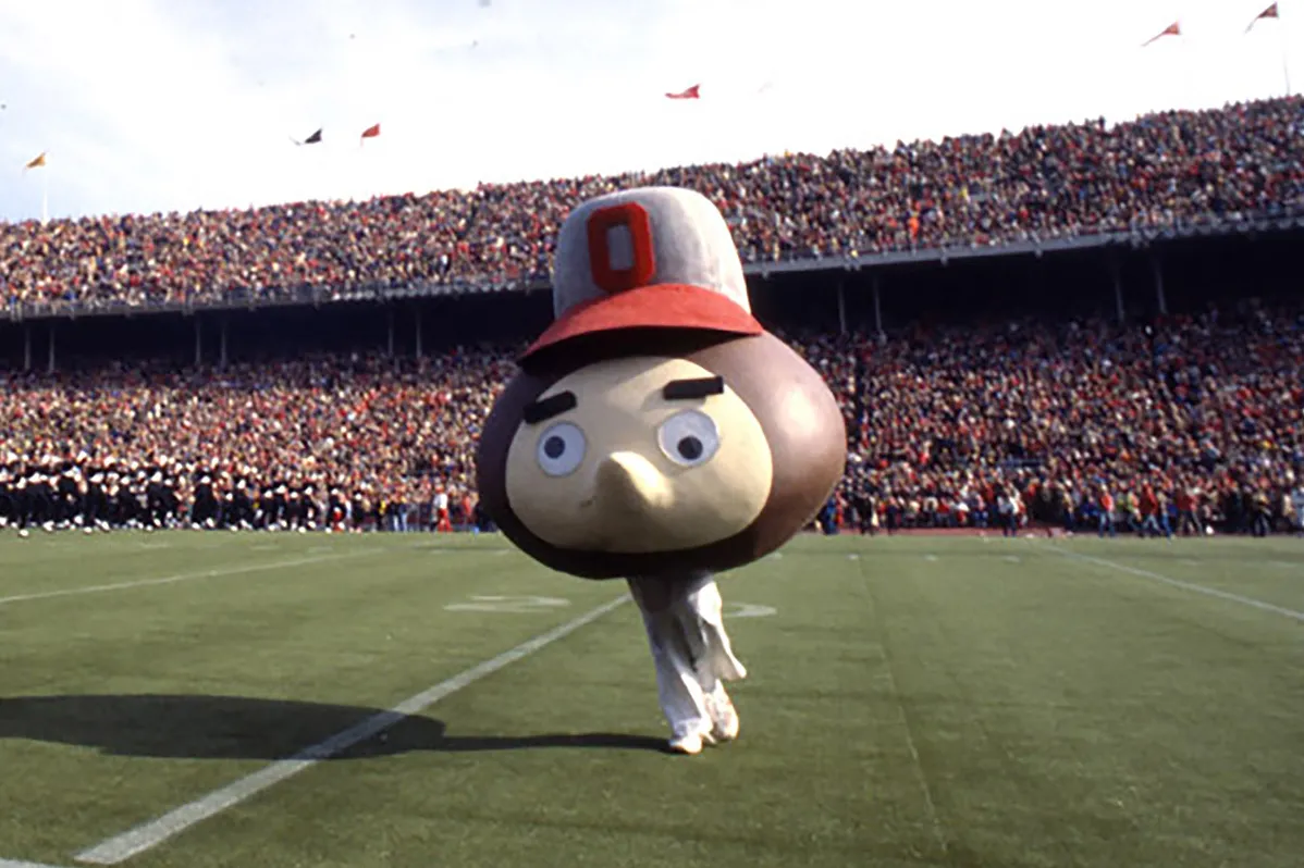 Another early giant-headed Brutus Buckeye runs across the field in Ohio Stadium. The Brutus wears a gigantic ballcap and its eyeballs point in different directions. It has a large nose, but no mouth, and the head comes down to the student's hips. The over-all impression is kind of weird.
