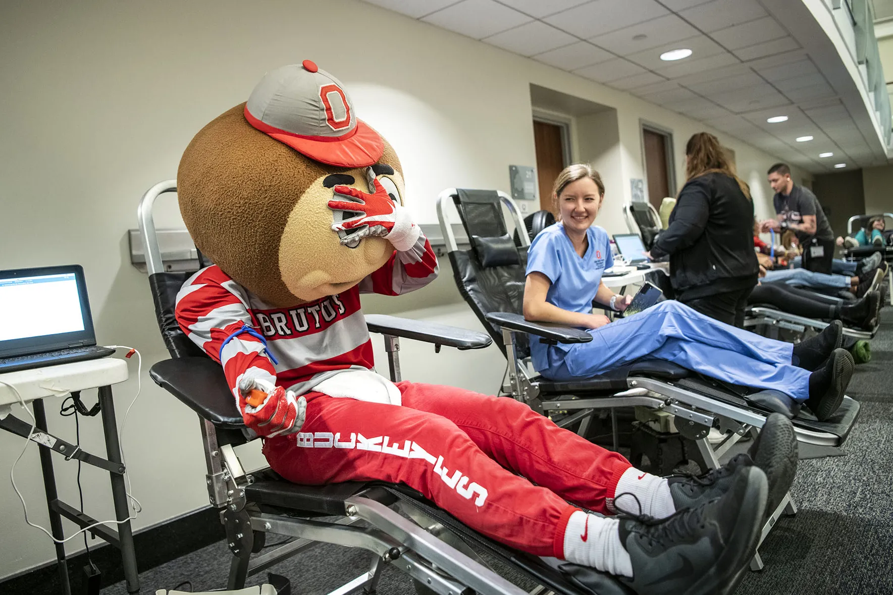 Brutus covers his face while sitting in a donor's chair at a blood donation drive. His arm is outstretched like he's ready for the needle.